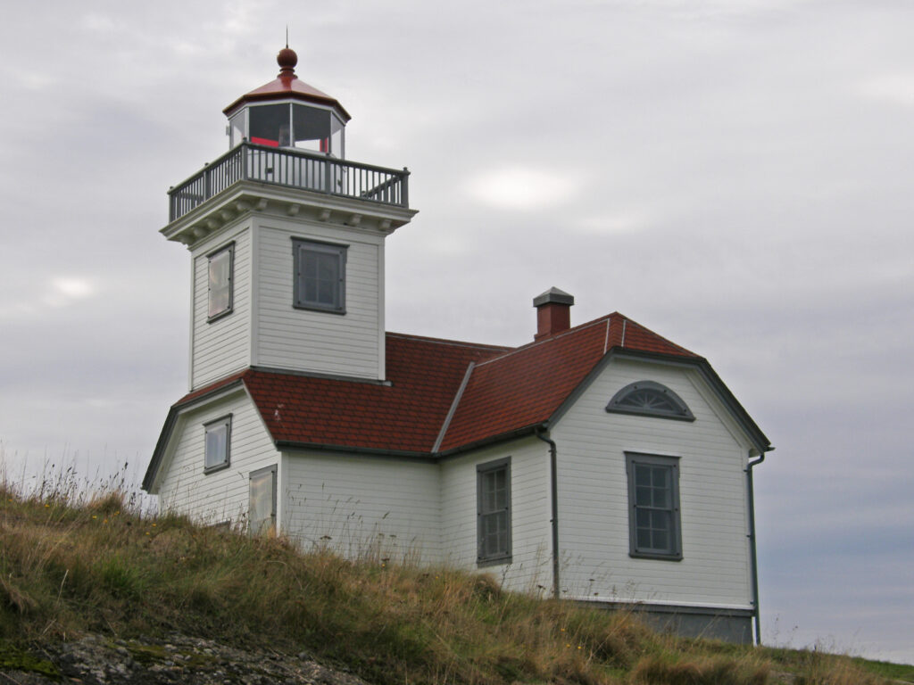 Lighthouse building with white walls and red roofing. Overcast gray sky with clouds. Lighthouse sits on a grassy hill.