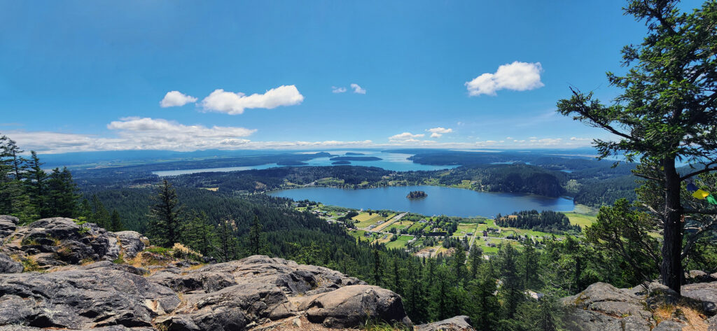 San Juan Island from a high vantage point overlooking a green landscape with forests, small settlements, and a large body of water with several small islands. In the distance, there are more islands and a blue sky with scattered white clouds. Trees frame the scene on both sides.