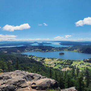 San Juan Island from a high vantage point overlooking a green landscape with forests, small settlements, and a large body of water with several small islands. In the distance, there are more islands and a blue sky with scattered white clouds. Trees frame the scene on both sides.