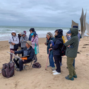 On the Normandy France beach, site of the World War 2 memorial. A group of watercolor artists standing on the sandy beach around a painter with an easel holding his paint brush. Its a cloudy and brisk day. Painter is wearing a large brim hat to protect from the weather and sun. His jacket is blue matching his pants.