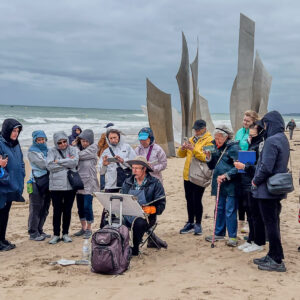 On the Normandy France beach, site of the World War 2 memorial. A group of watercolor artists standing on the sandy beach around a painter with an easel holding his paint brush. Its a cloudy and brisk day. Painter is wearing a large brim hat to protect from the weather and sun. His jacket is blue matching his pants.