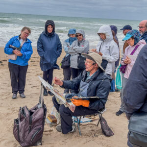 On the Normandy France beach, site of the World War 2 memorial. A group of watercolor artists standing on the sandy beach around a painter with an easel holding his paint brush. Its a cloudy and brisk day. Painter is wearing a large brim hat to protect from the weather and sun. His jacket is blue matching his pants.