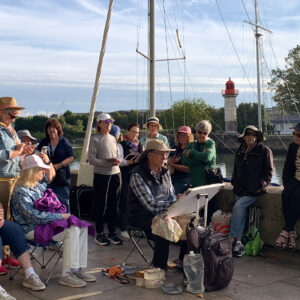 On a cobblestone walkway near the harbor in Honfluer France, a group of watercolor artists standing around a painter with an easel holding his paint brush. Painter is wearing a large brim hat to protect from the weather and sun.