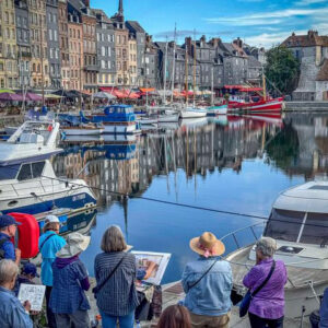 On a cobblestone walkway near the harbor in Honfluer France, a group of watercolor artists standing around a painter with an easel holding his paint brush. Painter is wearing a large brim hat to protect from the weather and sun.