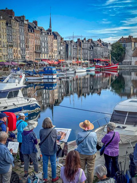 On a cobblestone walkway near the harbor in Honfluer France, a group of watercolor artists standing around a painter with an easel holding his paint brush. Painter is wearing a large brim hat to protect from the weather and sun.