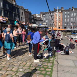 On a cobblestone walkway near the harbor in Honfluer France, a group of watercolor artists standing around a painter with an easel holding his paint brush. Painter is wearing a large brim hat to protect from the weather and sun.