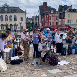 On a cobblestone walkway near the harbor in Honfluer France, a group of watercolor artists standing around a painter with an easel holding his paint brush. Painter is wearing a large brim hat to protect from the weather and sun.