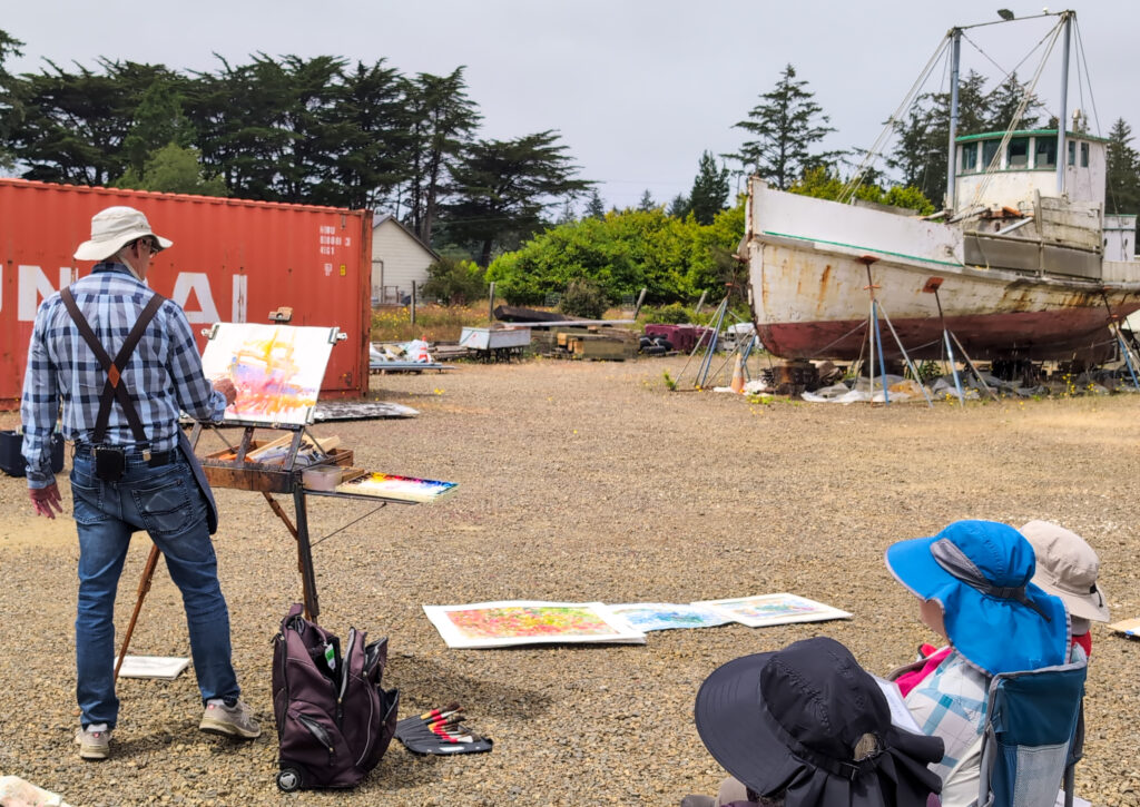 Watercolor artist standing in front of an easel which is distantly placed in front of an old ship in the yard for repair.