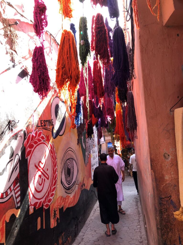 Photo reference of a Moroccan market place with people on a busy street and images painted on the walls and wares suspended from above.