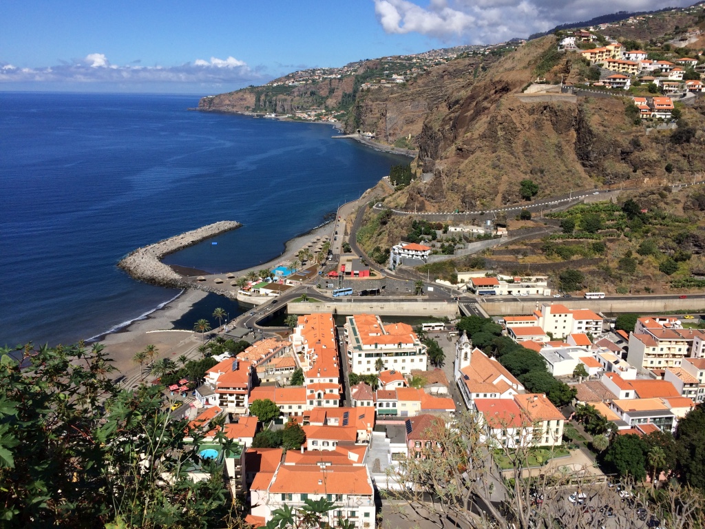 Mountain cliffs over a small coastal town where the houses all have terra cotta roofs. 